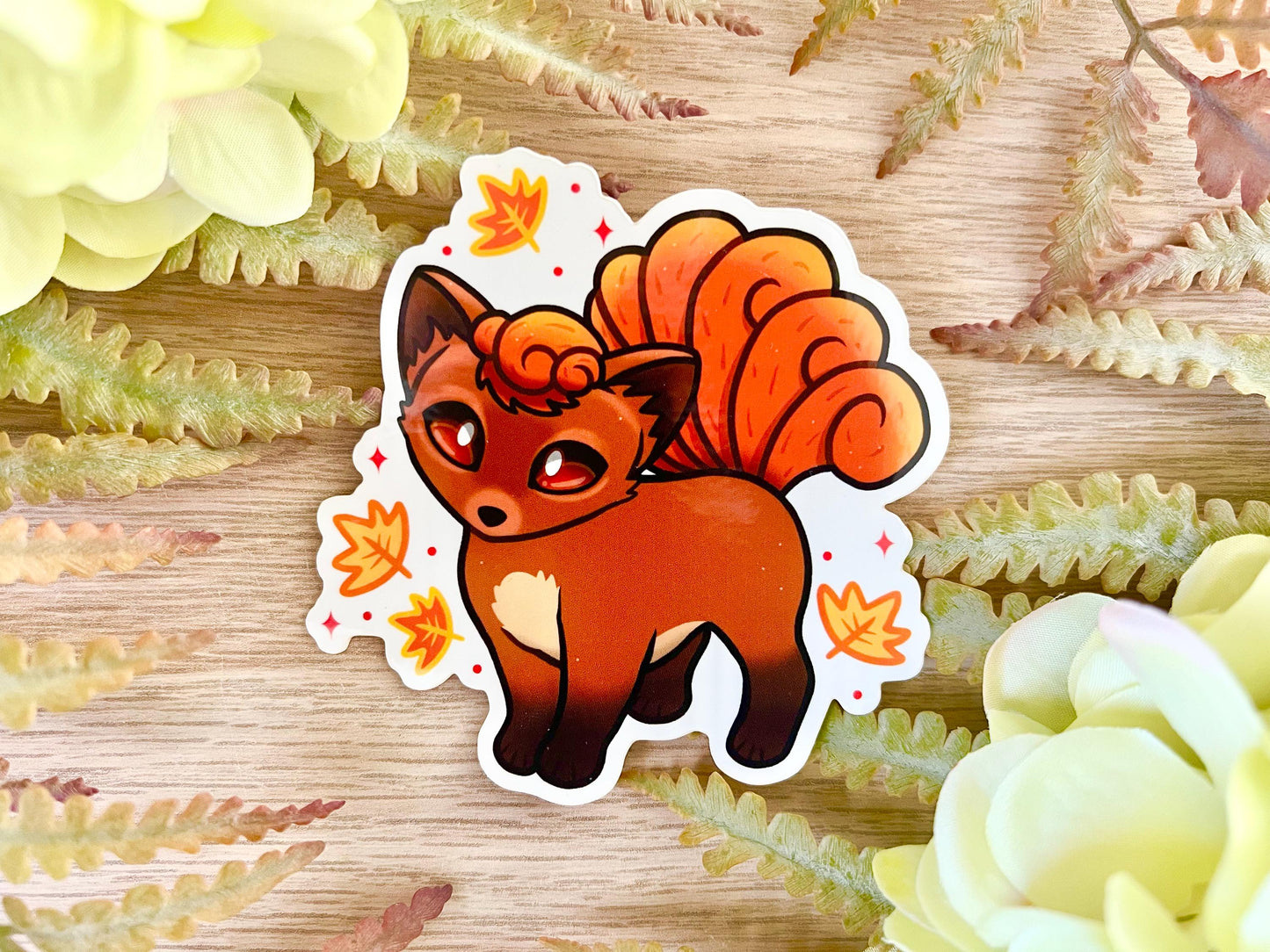 A sticker of a cute orange fox creature with 6 tails and big brown eyes surrounded by orange and yellow leaves.