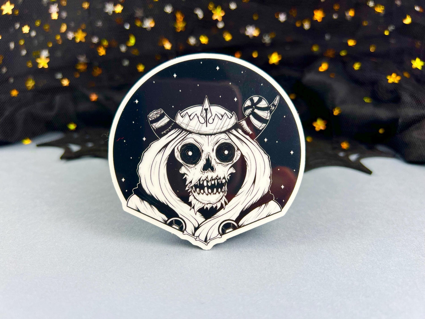 A glow in the dark black and white sticker of a skull with horns, a crown, a hood, and cape in front of a background of a dark starry sky.