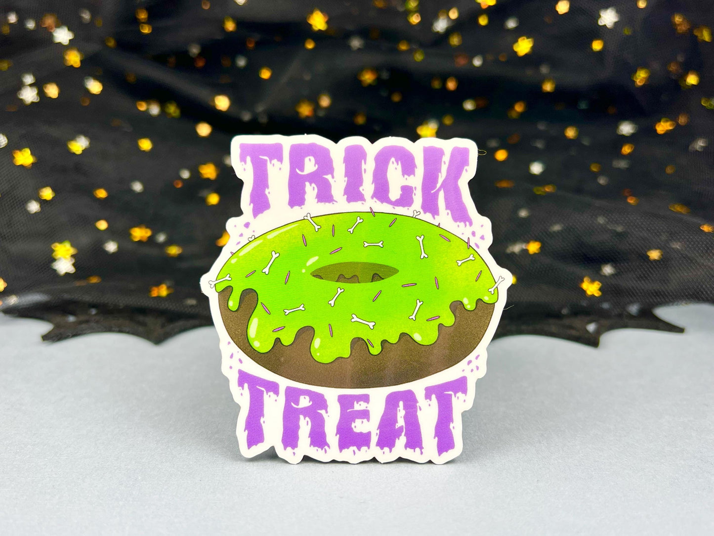 A deliciously spooky transparent donut sticker with green frosting, bone and purple sprinkles, and purple trick or treat writing on the top and bottom.