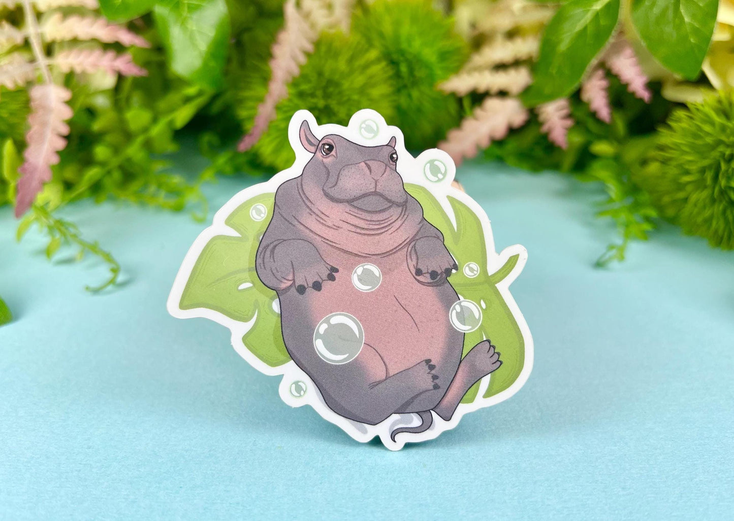 A sticker of a cute hippo laying on its back on top of a monstera leaf playing with bubbles.