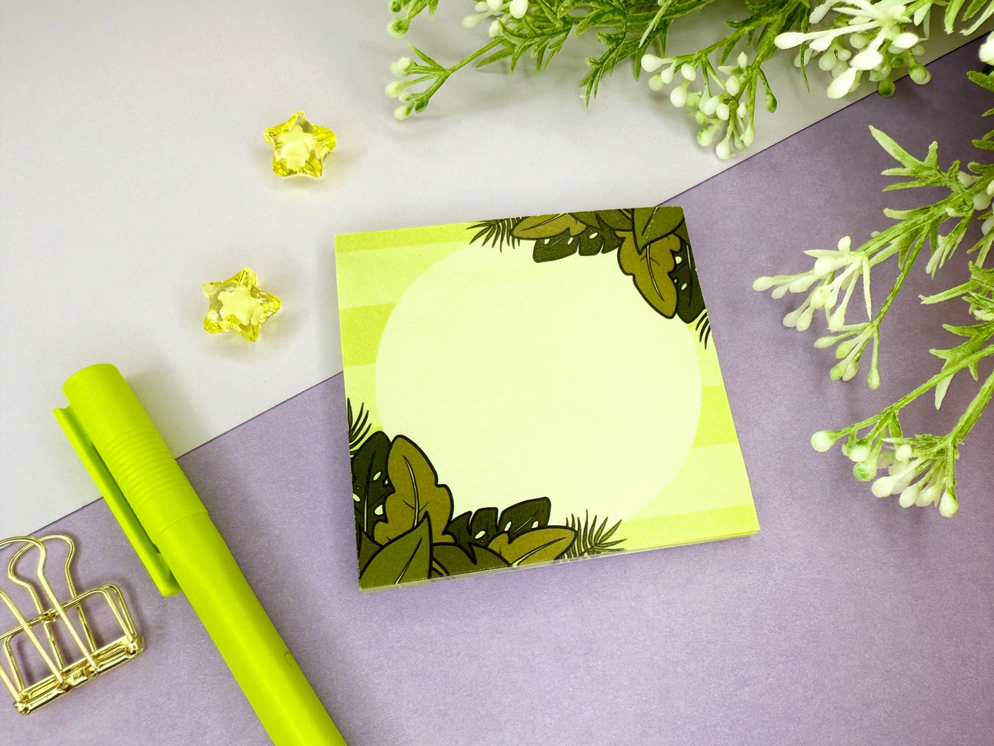Green sticky note with a tropical leaf design on a purple surface with flowers and a green pen.