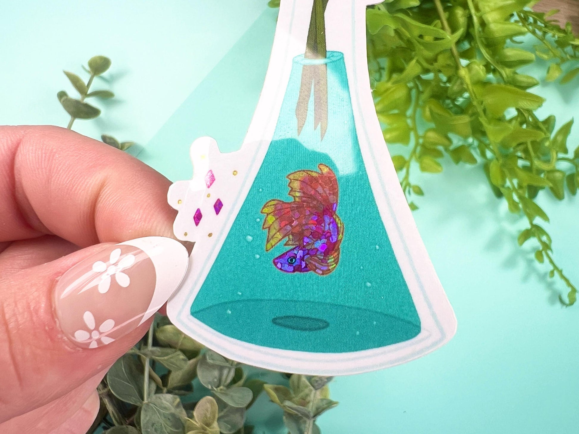 A vase filled with pink and yellow glitter Snapdragon flowers and has water with a pink, orange, and yellow glitter Betta fish inside.