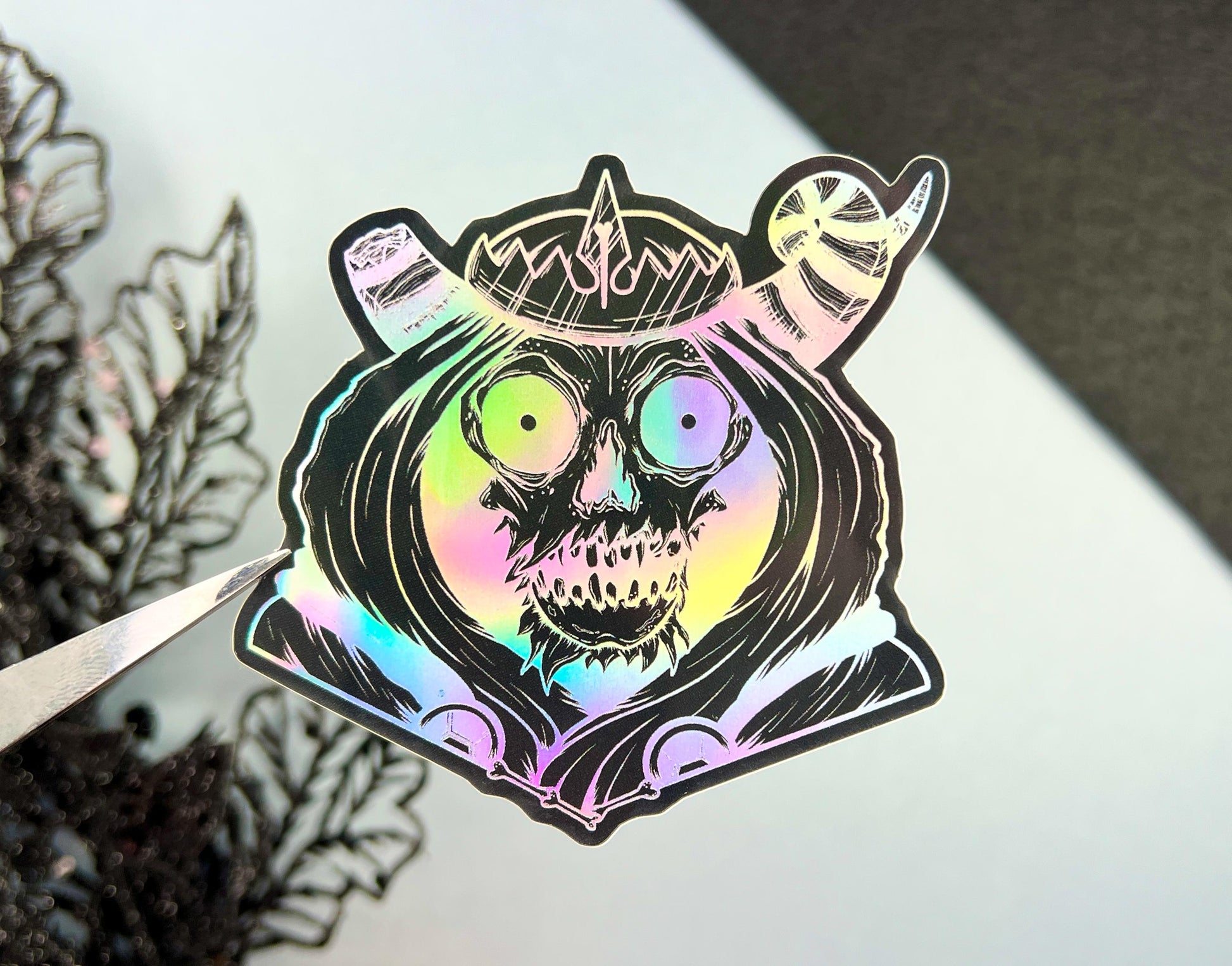 A black and silver holographic sticker of a skull with horns, a crown, a hood, and cape.