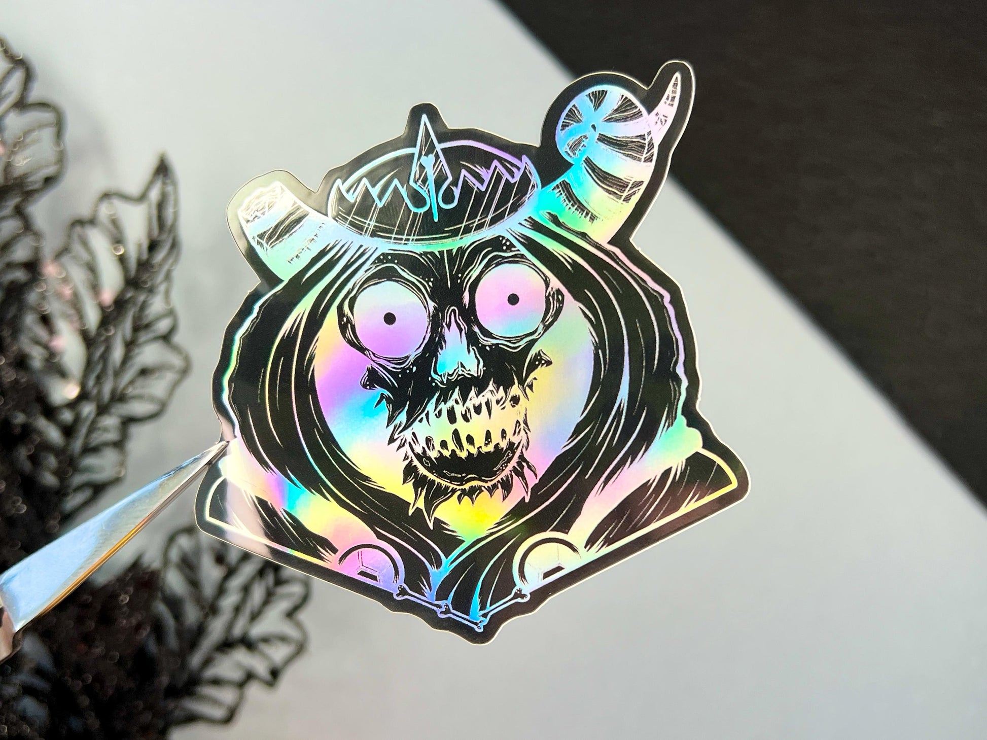 A black and silver holographic sticker of a skull with horns, a crown, a hood, and cape.