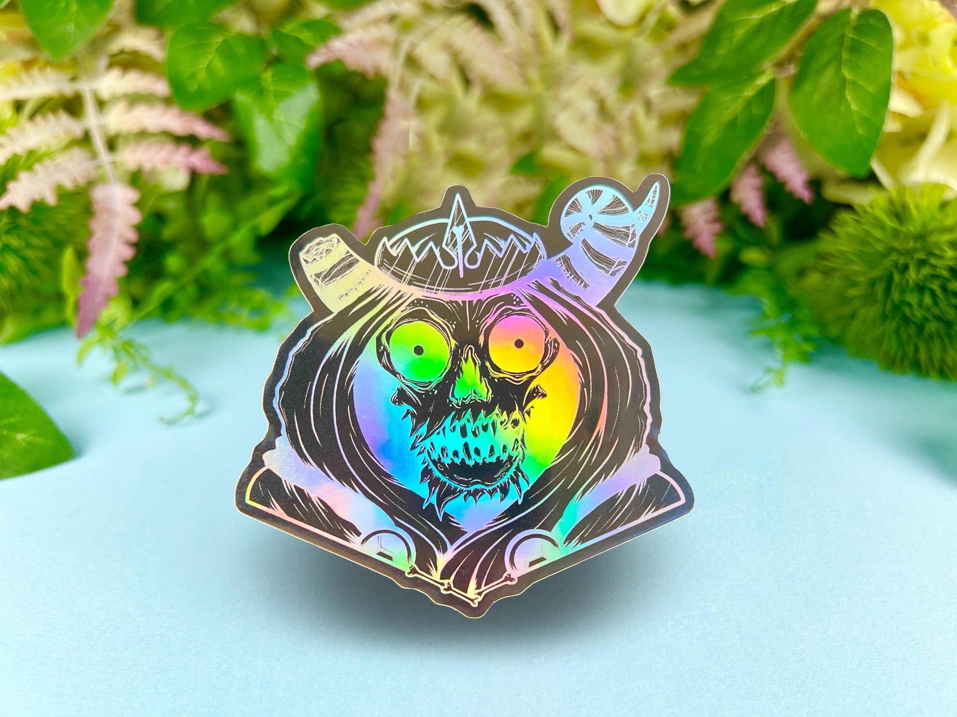 A black and silver holographic sticker of a skull with horns, a crown, a hood, and cape.