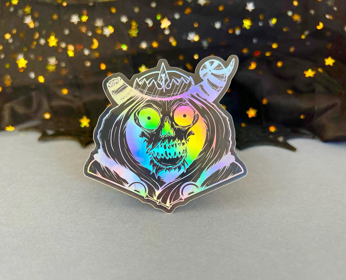 A black and silver holographic sticker of a skull with horns, a crown, a hood, and cape.