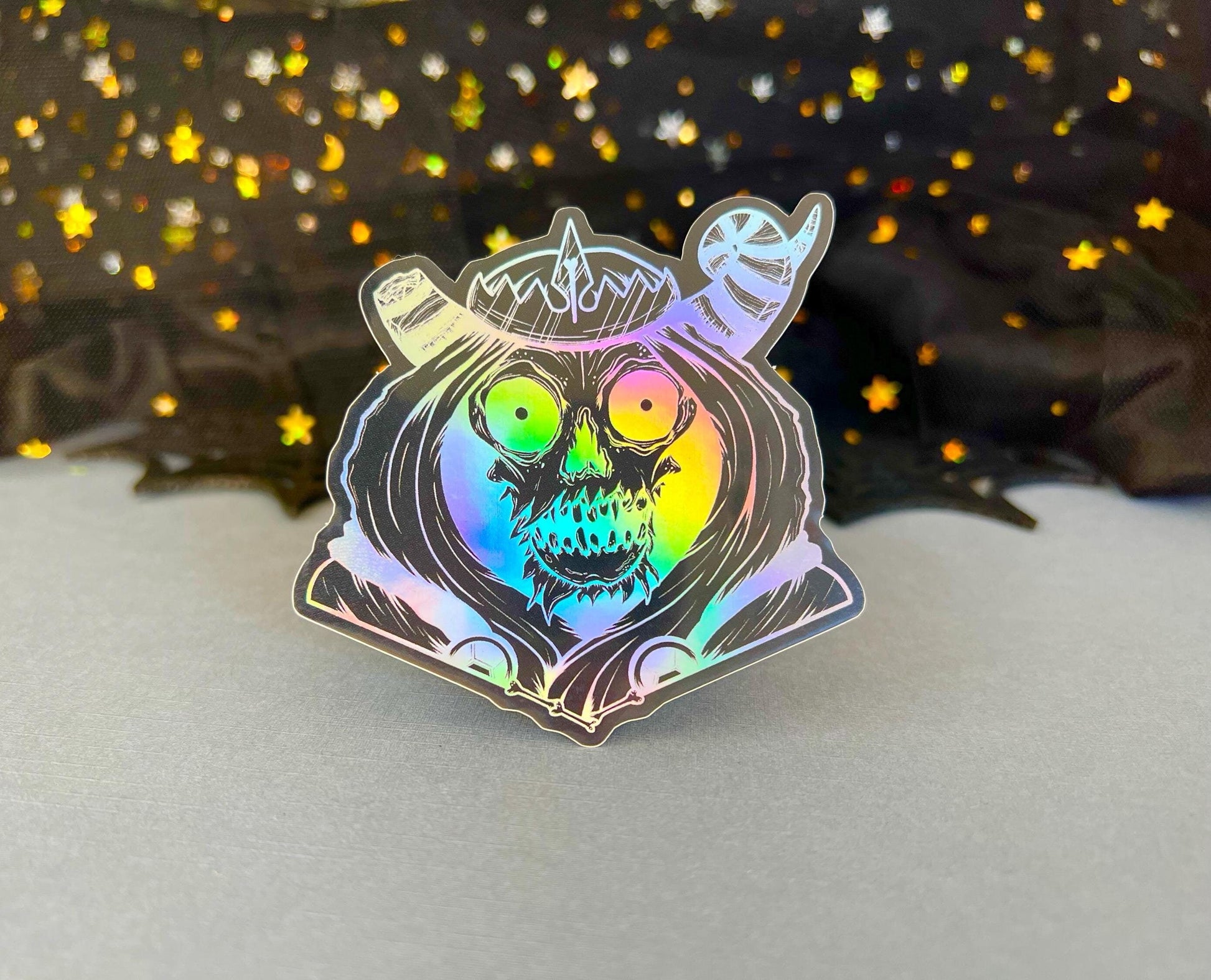 A black and silver holographic sticker of a skull with horns, a crown, a hood, and cape.