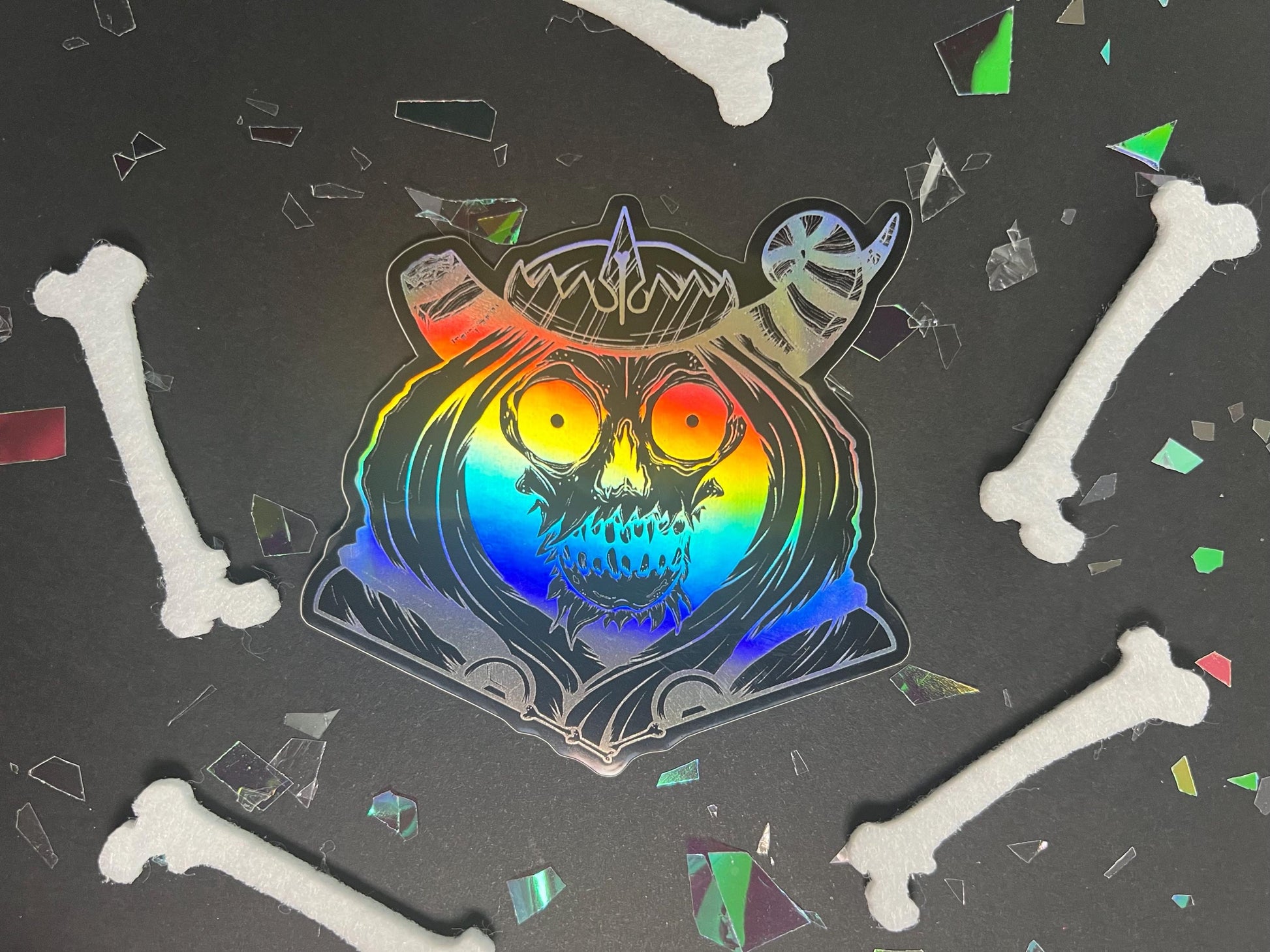 A black and silver holographic sticker of a skull with horns, a crown, a hood, and cape.