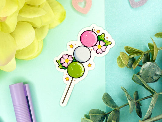 Pink, white, and green dango mochi on a stick with pink blossoms around it.