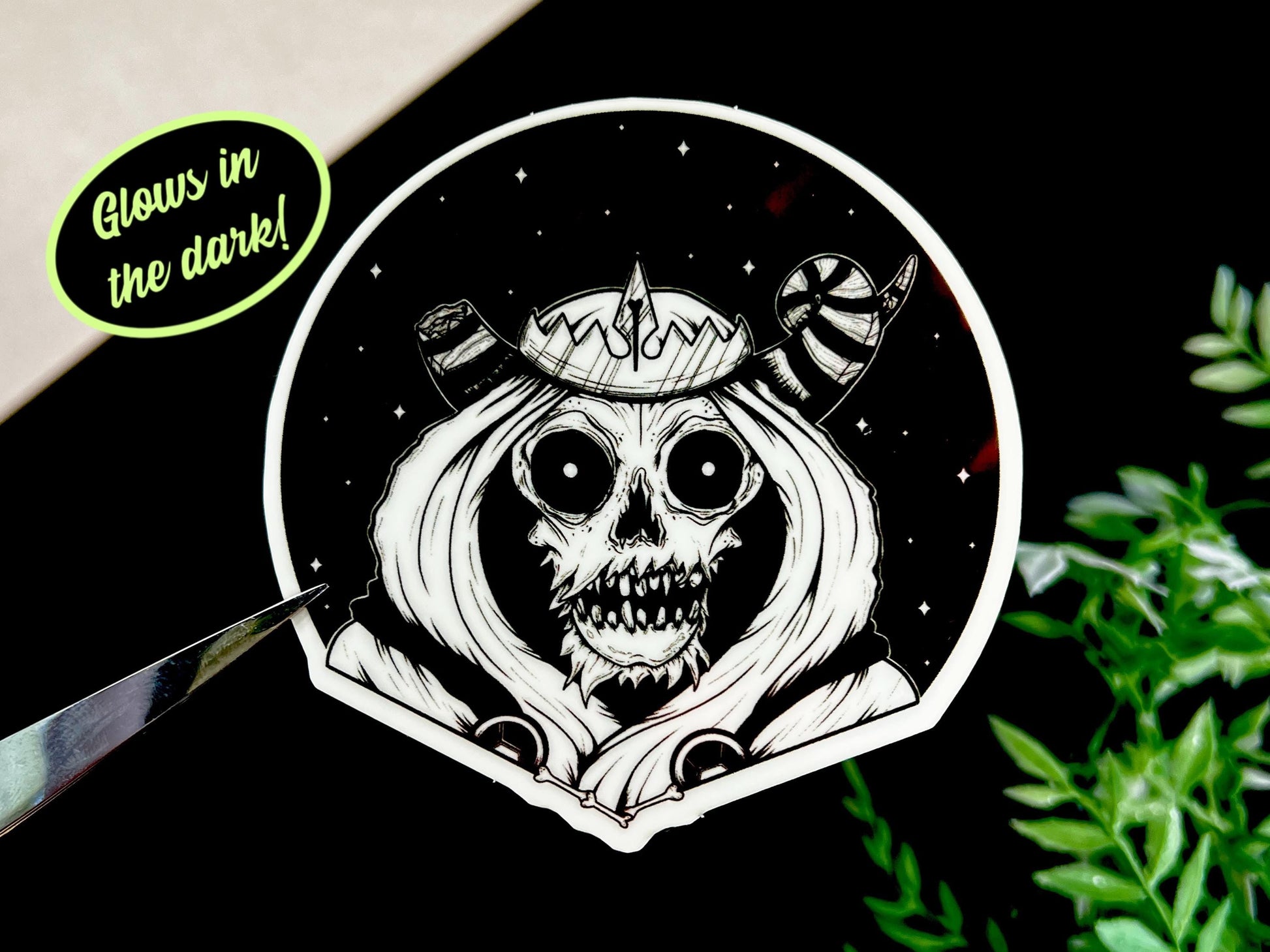 A glow in the dark black and white sticker of a skull with horns, a crown, a hood, and cape in front of a background of a dark starry sky.