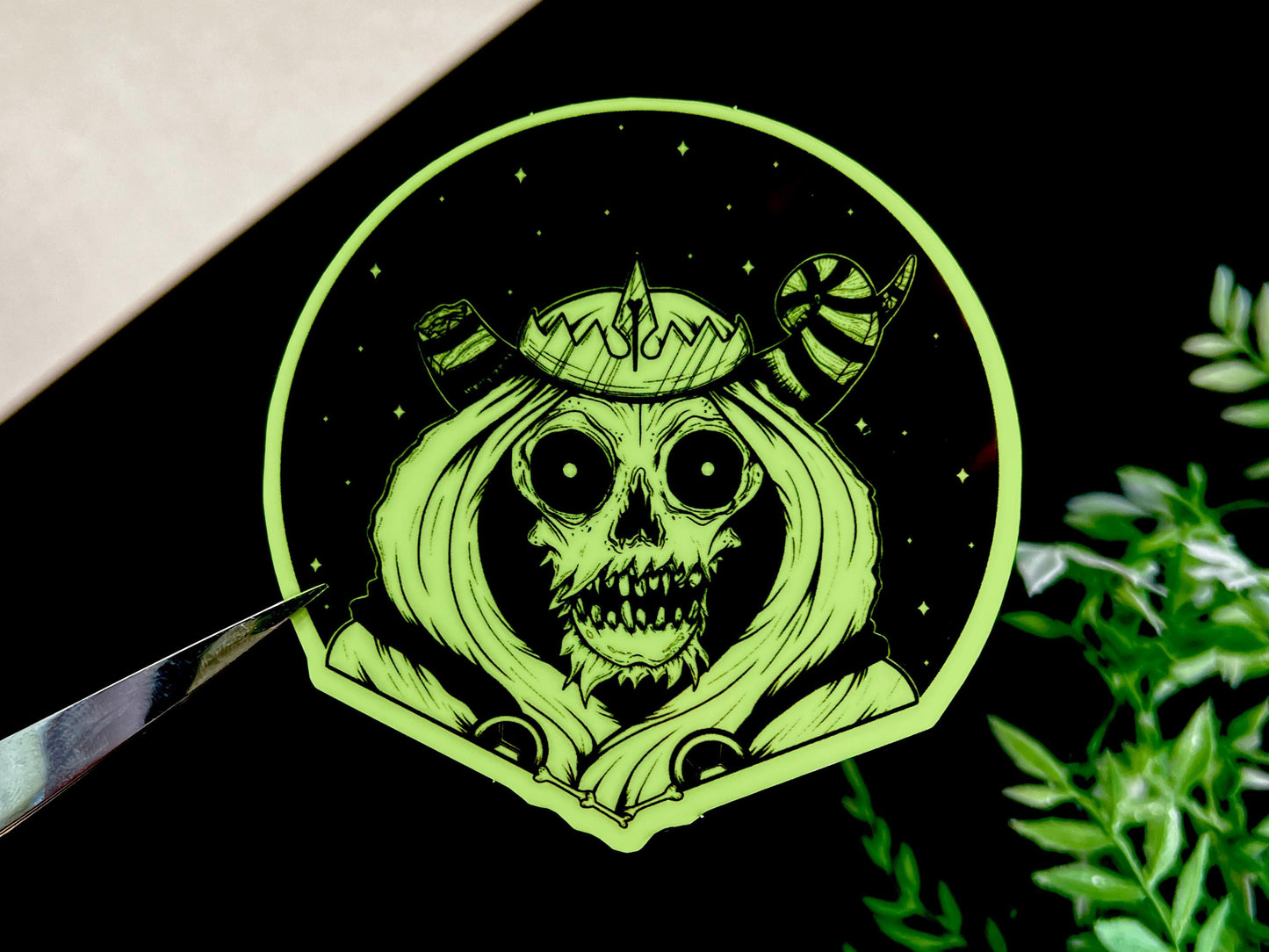 A glow in the dark black and white sticker of a skull with horns, a crown, a hood, and cape in front of a background of a dark starry sky.