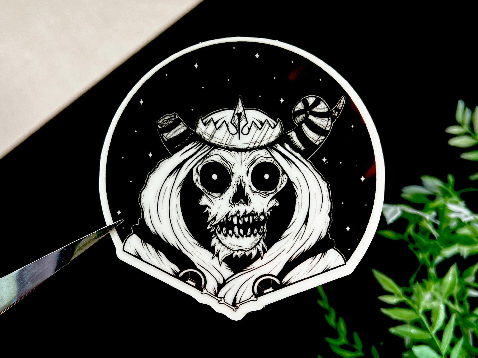 A glow in the dark black and white sticker of a skull with horns, a crown, a hood, and cape in front of a background of a dark starry sky.