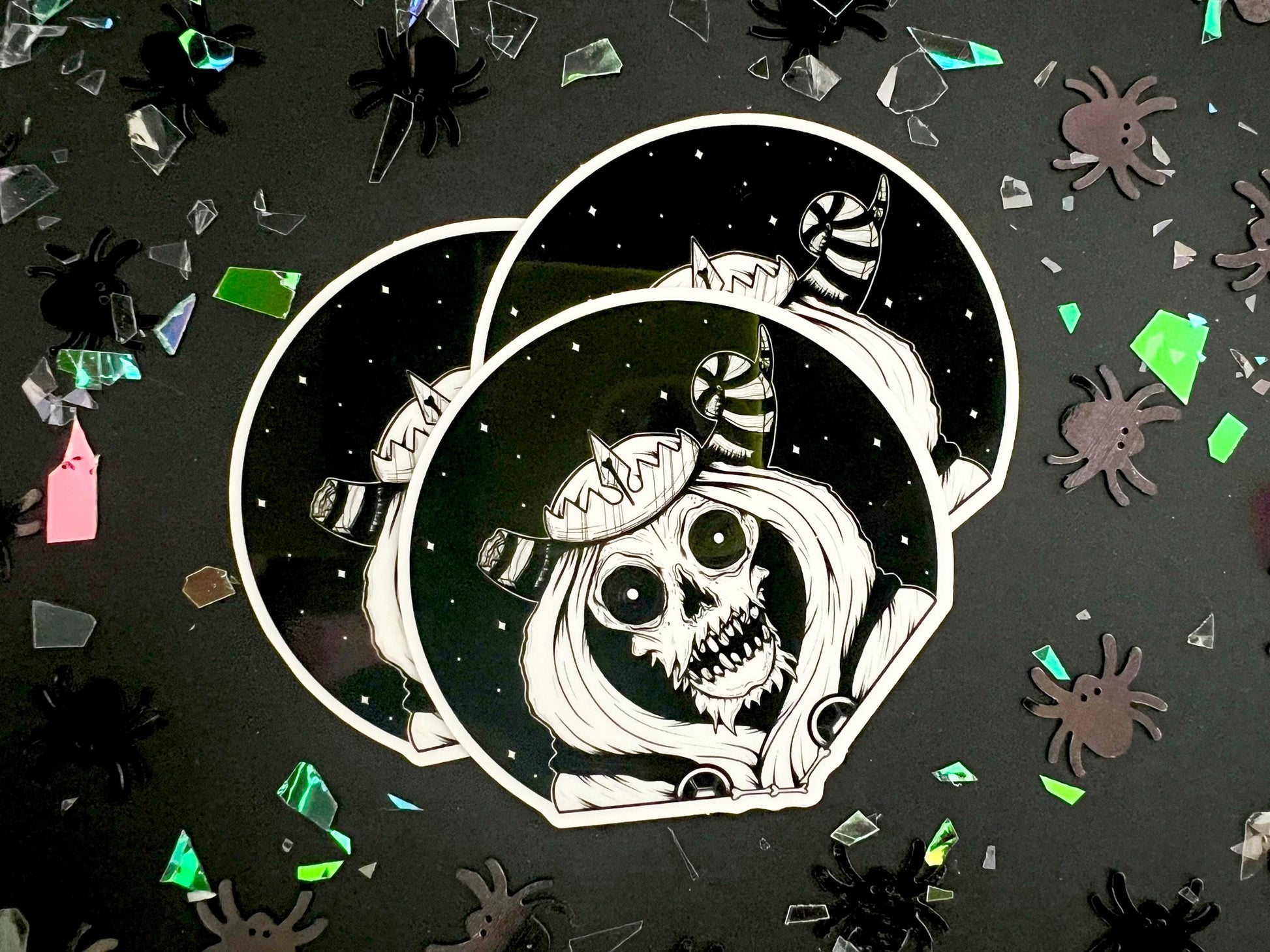 A glow in the dark black and white sticker of a skull with horns, a crown, a hood, and cape in front of a background of a dark starry sky.