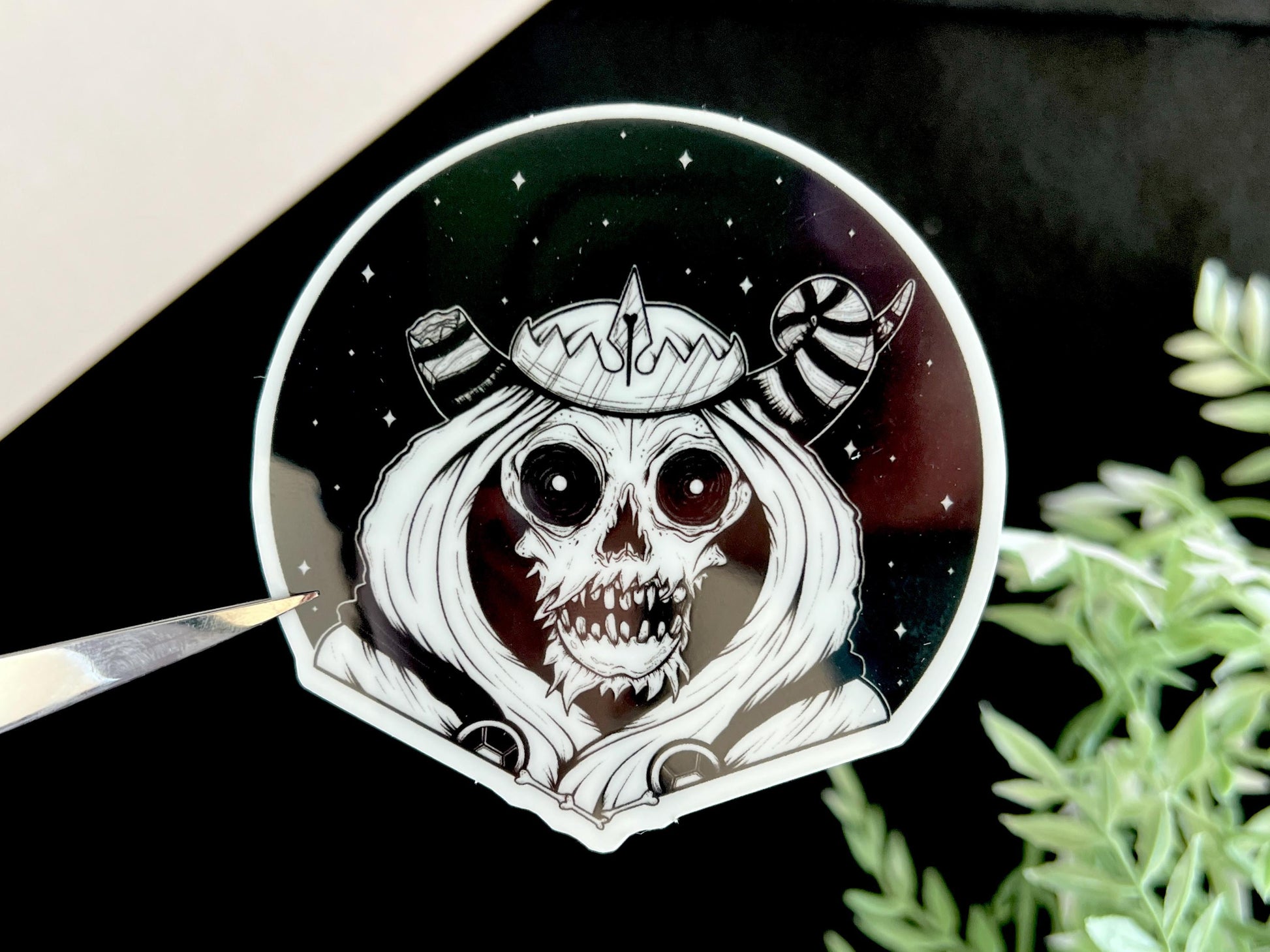 A glow in the dark black and white sticker of a skull with horns, a crown, a hood, and cape in front of a background of a dark starry sky.
