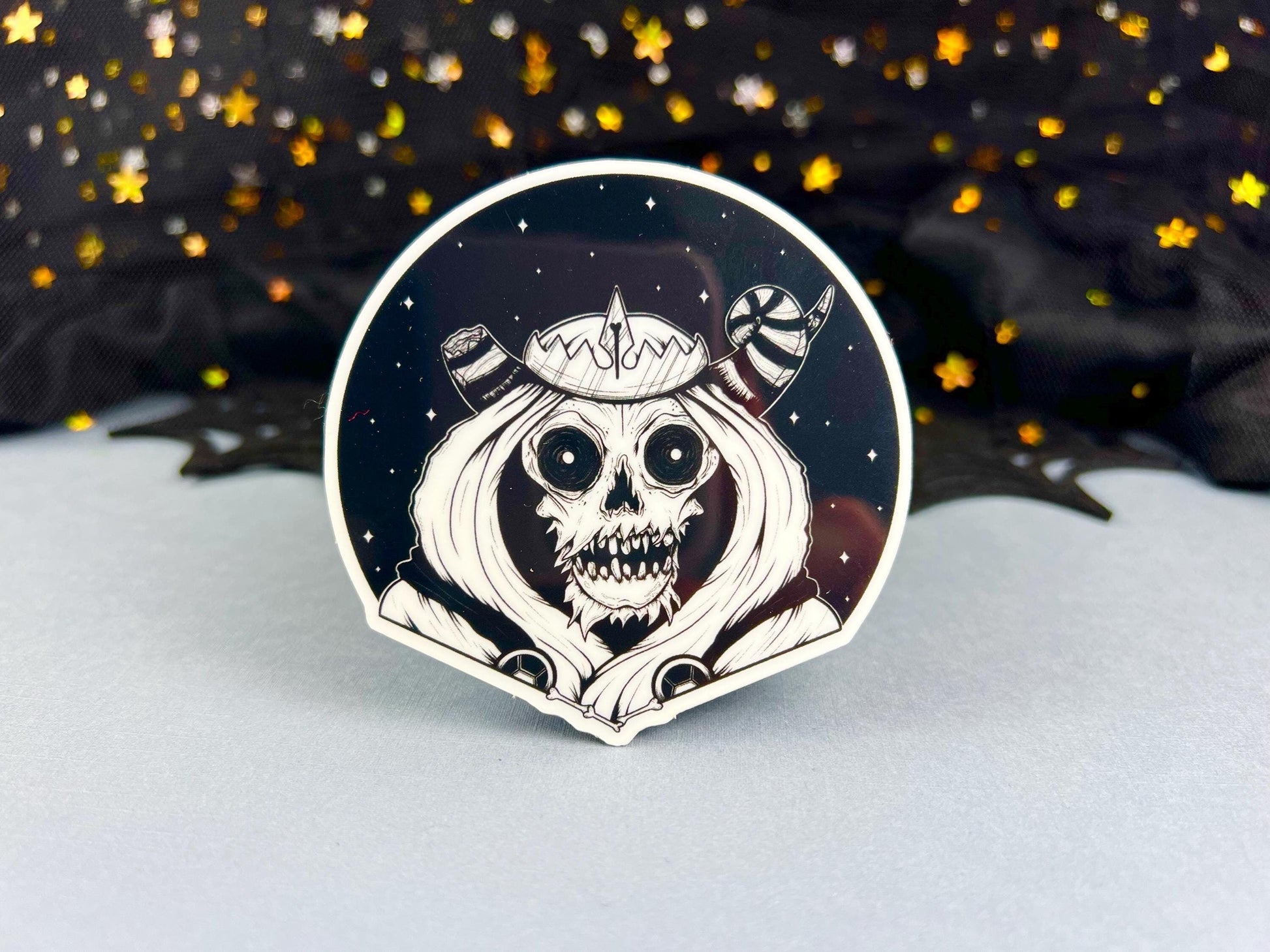 A glow in the dark black and white sticker of a skull with horns, a crown, a hood, and cape in front of a background of a dark starry sky.