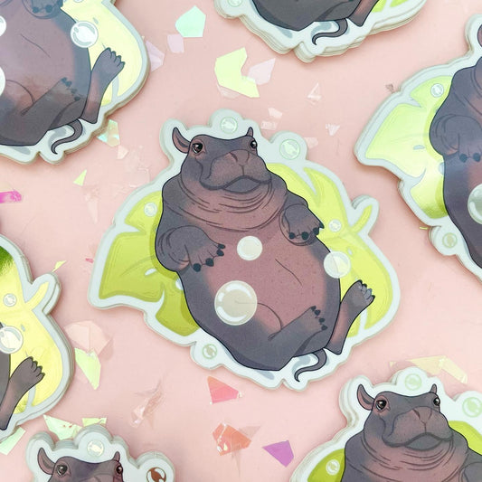 A sticker of a cute hippo laying on its back on top of a monstera leaf playing with bubbles.
