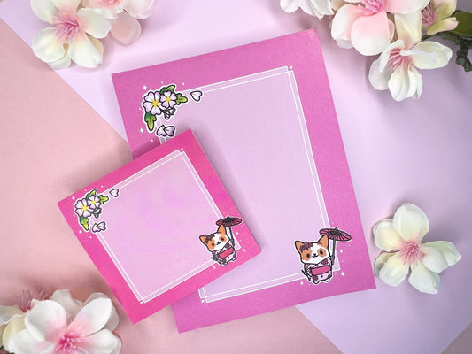 A pink gift set, including sticky notes and a notepad, that has an illustration of a cute dog wearing a pink kimono holding an umbrella under cherry blossoms. 