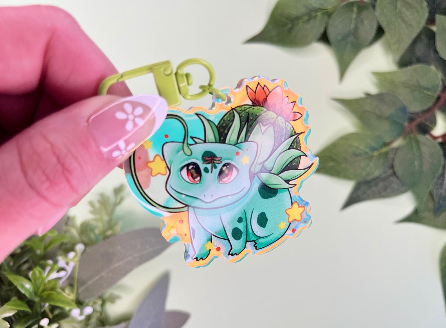 A keychain of a cute 4 legged green plant creature with a cactus and orange flower on its back, vines coming out their side , and yellow glowing stars surrounding them.