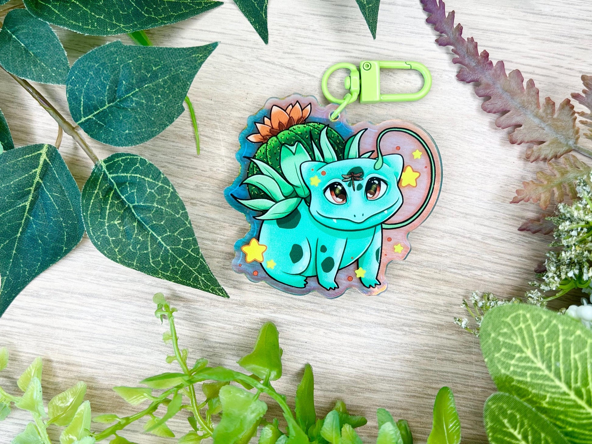 A keychain of a cute 4 legged green plant creature with a cactus and orange flower on its back, vines coming out their side , and yellow glowing stars surrounding them.