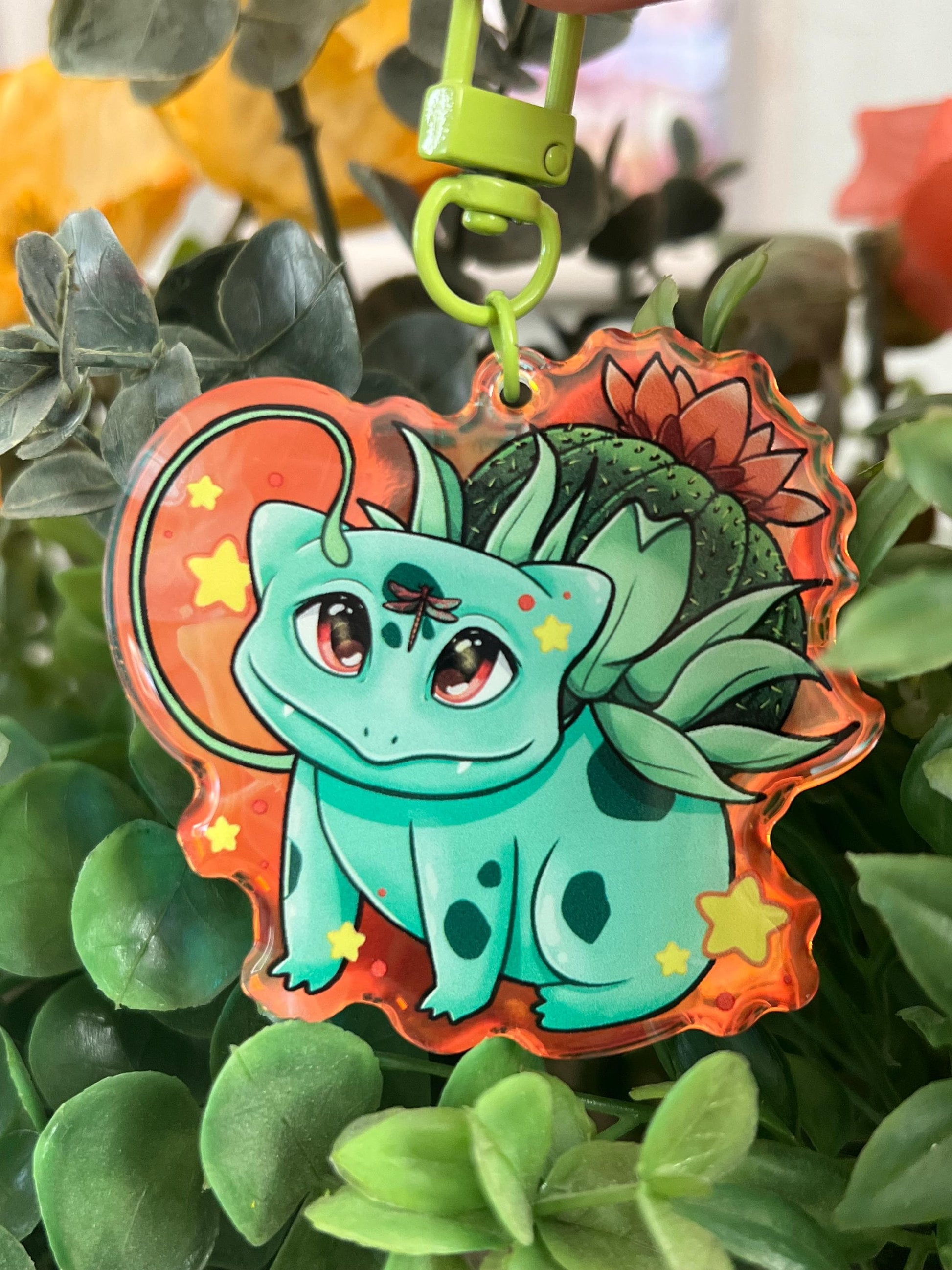 A keychain of a cute 4 legged green plant creature with a cactus and orange flower on its back, vines coming out their side , and yellow glowing stars surrounding them.