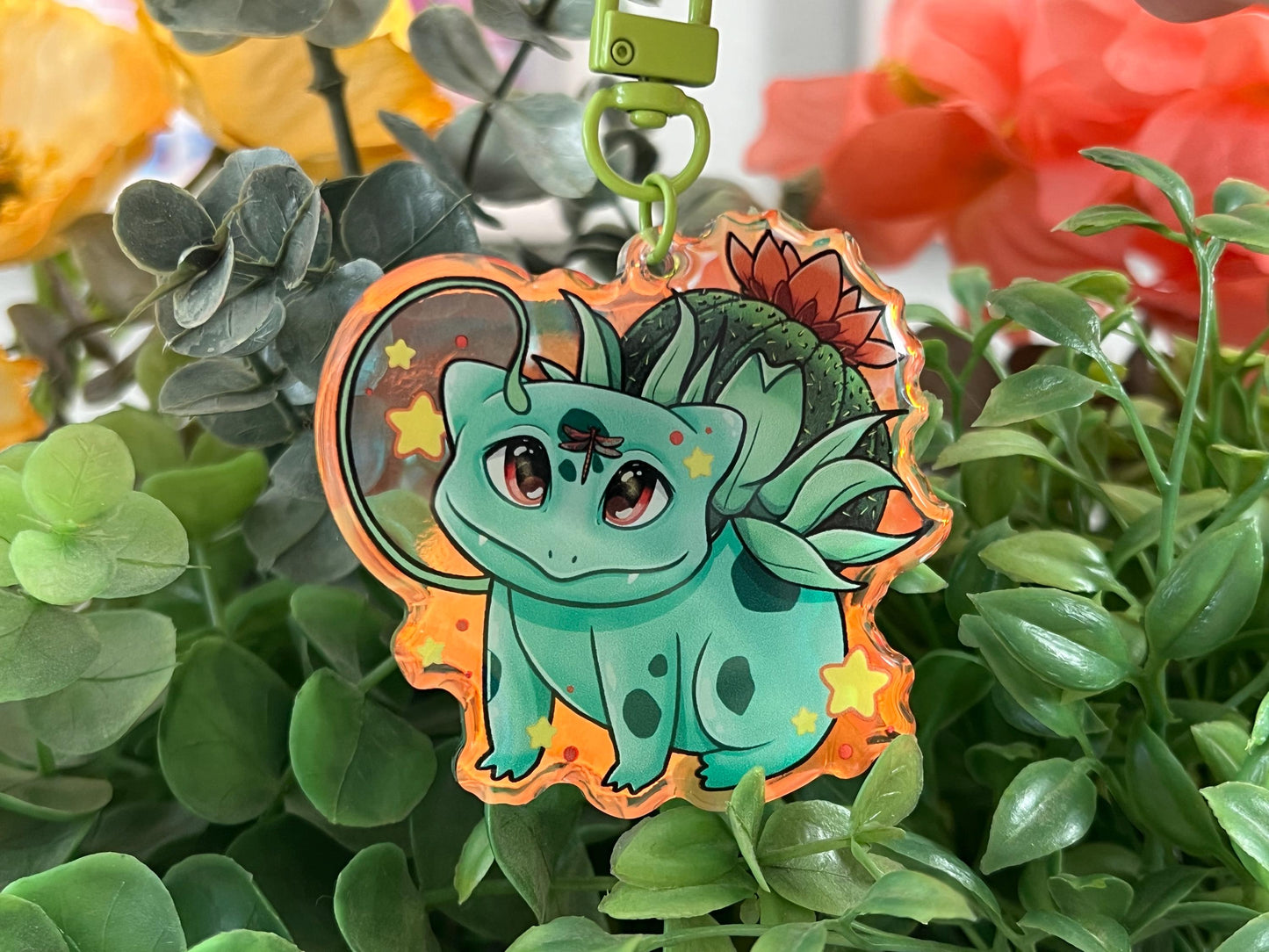 A keychain of a cute 4 legged green plant creature with a cactus and orange flower on its back, vines coming out their side , and yellow glowing stars surrounding them.