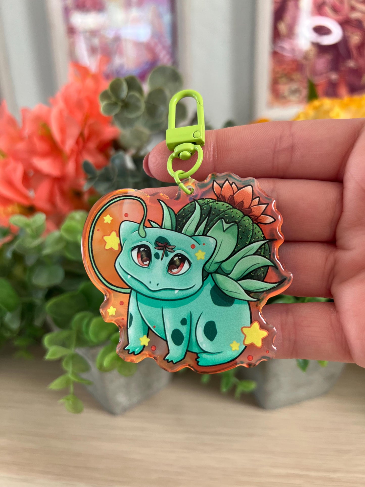 A keychain of a cute 4 legged green plant creature with a cactus and orange flower on its back, vines coming out their side , and yellow glowing stars surrounding them.