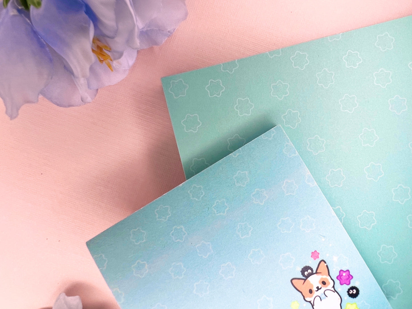 A blue gift set that includes one sticky note pad and one notepad with an illustration of a cute dog playing with soot sprites and rainbow konpieto and has a konpieto patterned background.