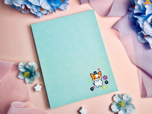 A blue notepad with an illustration of a cute dog playing with soot sprites and rainbow konpieto and has a konpieto patterned background.
