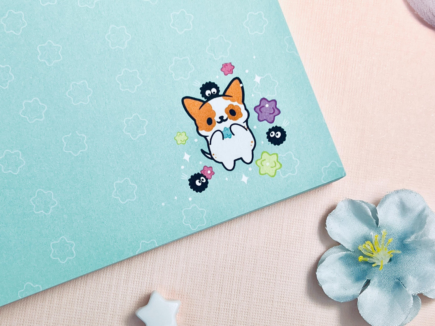 A blue notepad with an illustration of a cute dog playing with soot sprites and rainbow konpieto and has a konpieto patterned background.
