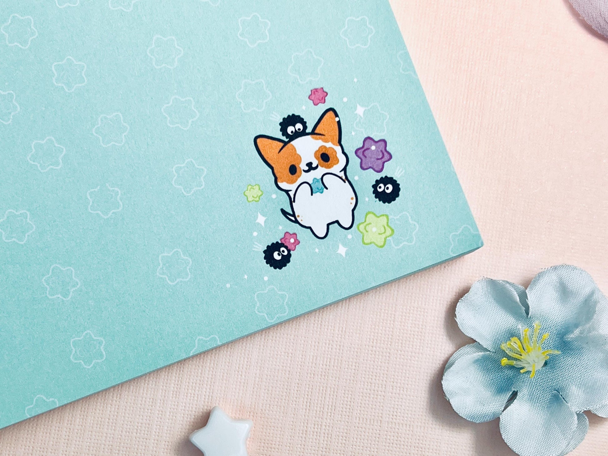 A blue notepad with an illustration of a cute dog playing with soot sprites and rainbow konpieto and has a konpieto patterned background.