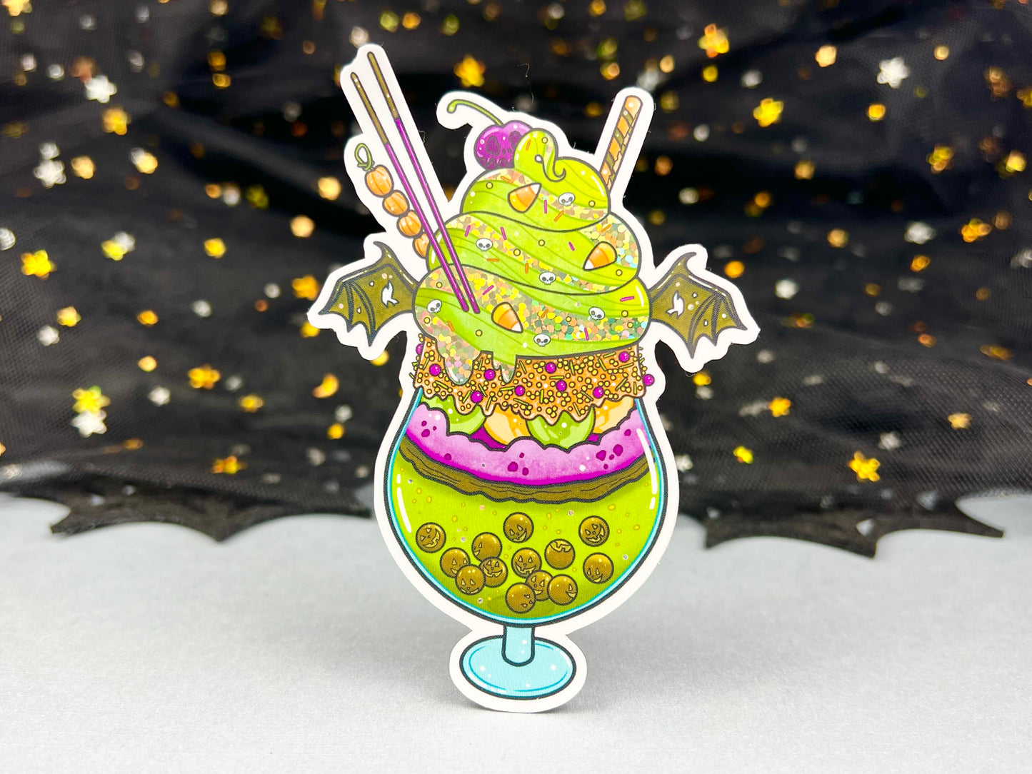 A green, orange, and purple sticker illustration of a spooky drink with jack o'lantern boba, frosting, sprinkles, whip cream, candy corn, a skull cherry, straw and bat wings.