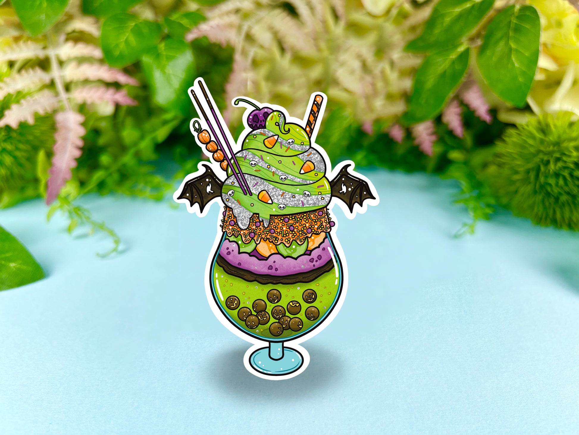 A green, orange, and purple sticker illustration of a spooky drink with jack o'lantern boba, frosting, sprinkles, whip cream, candy corn, a skull cherry, straw and bat wings.