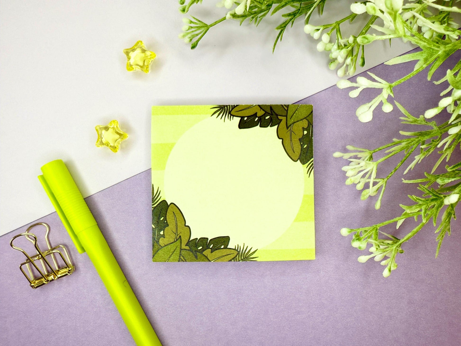 Green sticky note with a tropical leaf design on a purple surface with flowers and a green pen.