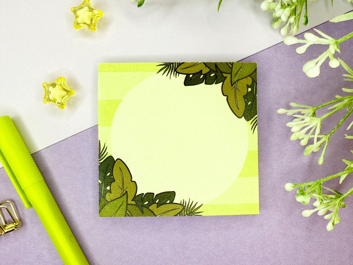 Green sticky note with a tropical leaf design on a purple surface with flowers and a green pen.