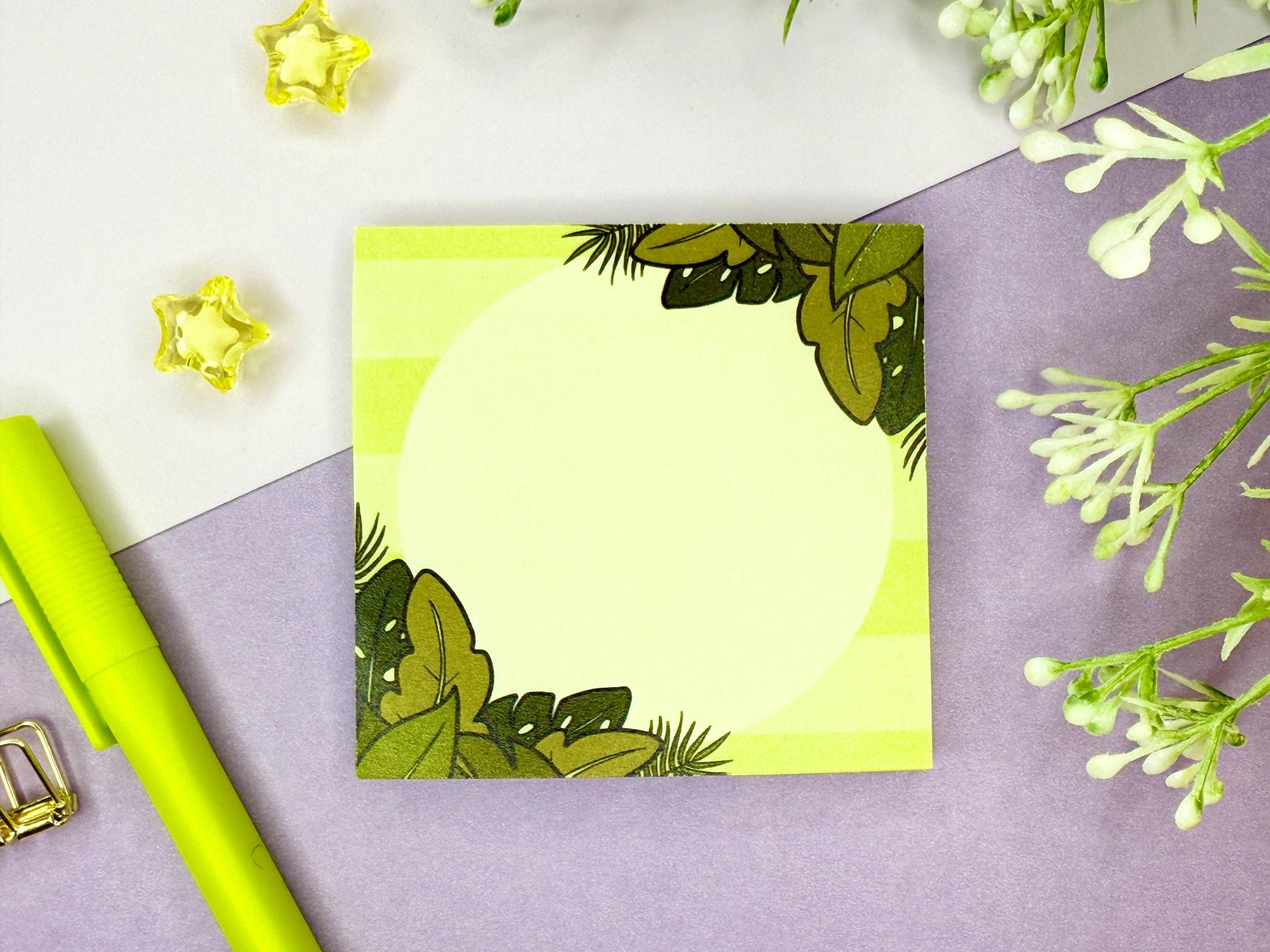 Green sticky note with a tropical leaf design on a purple surface with flowers and a green pen.
