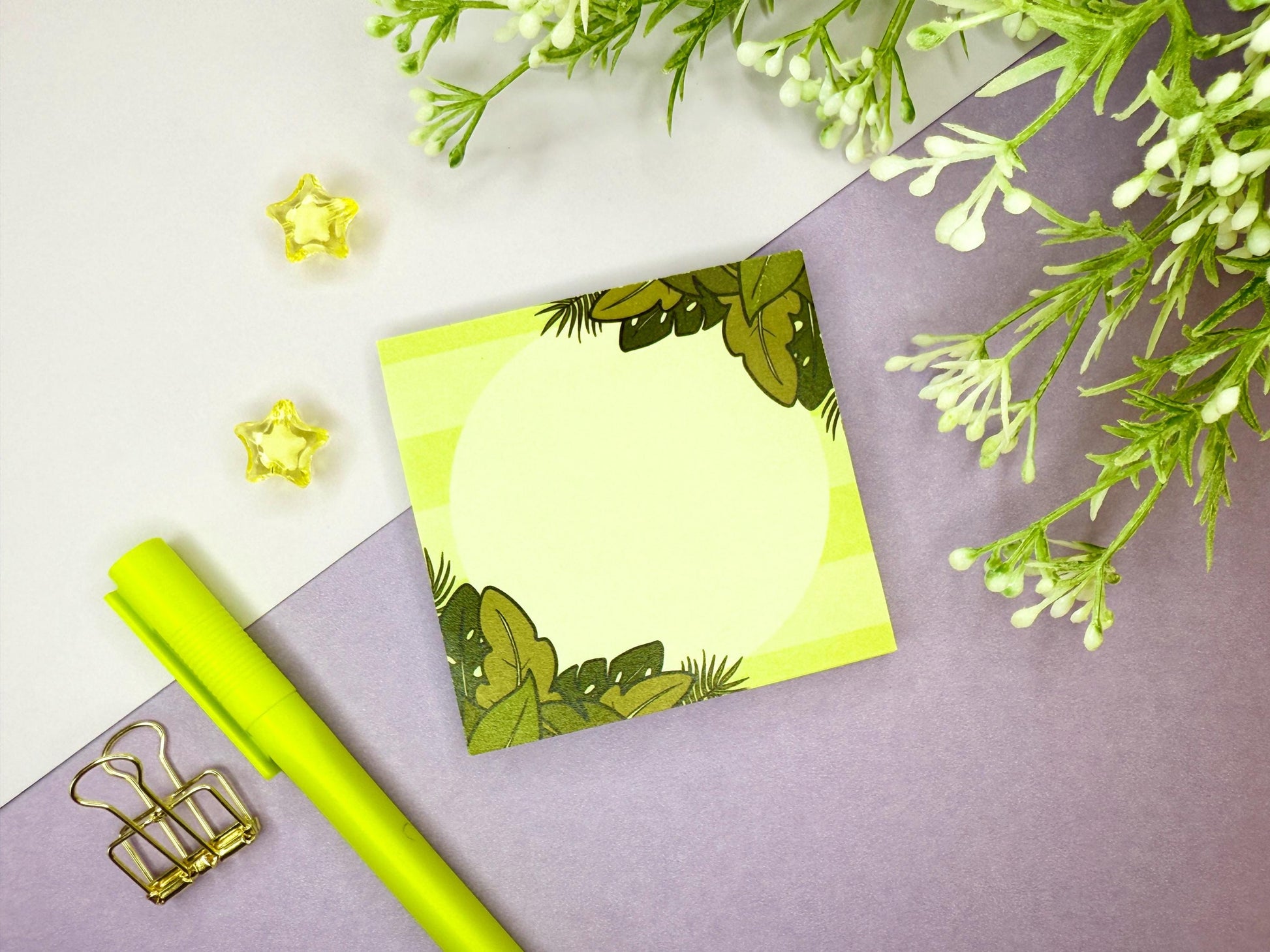 Green sticky note with a tropical leaf design on a purple surface with flowers and a green pen.