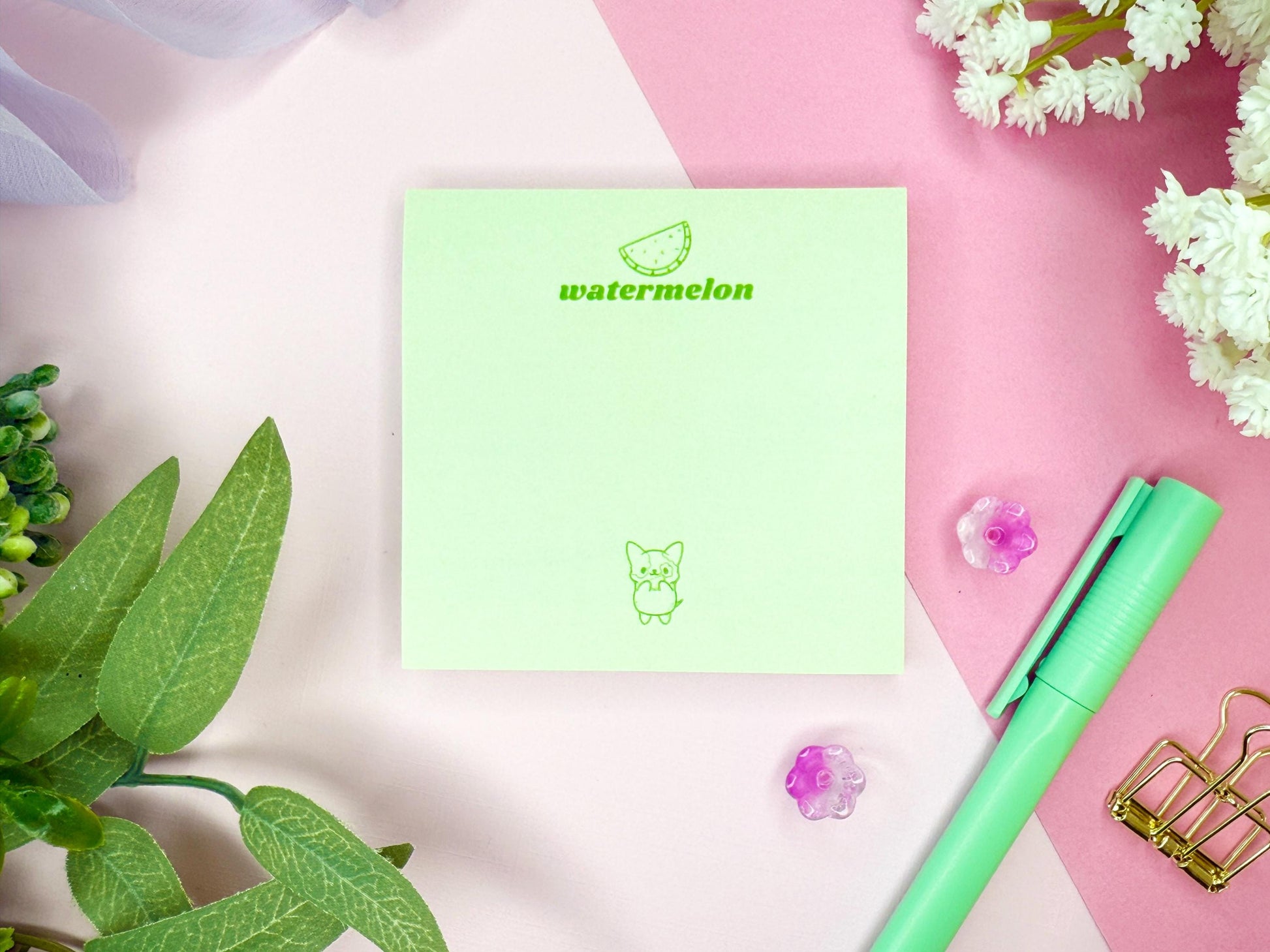 Light green notepad with "watermelon" branding and illustration as well as a kawaii dog with a green pen, and flowers on a pink and white background