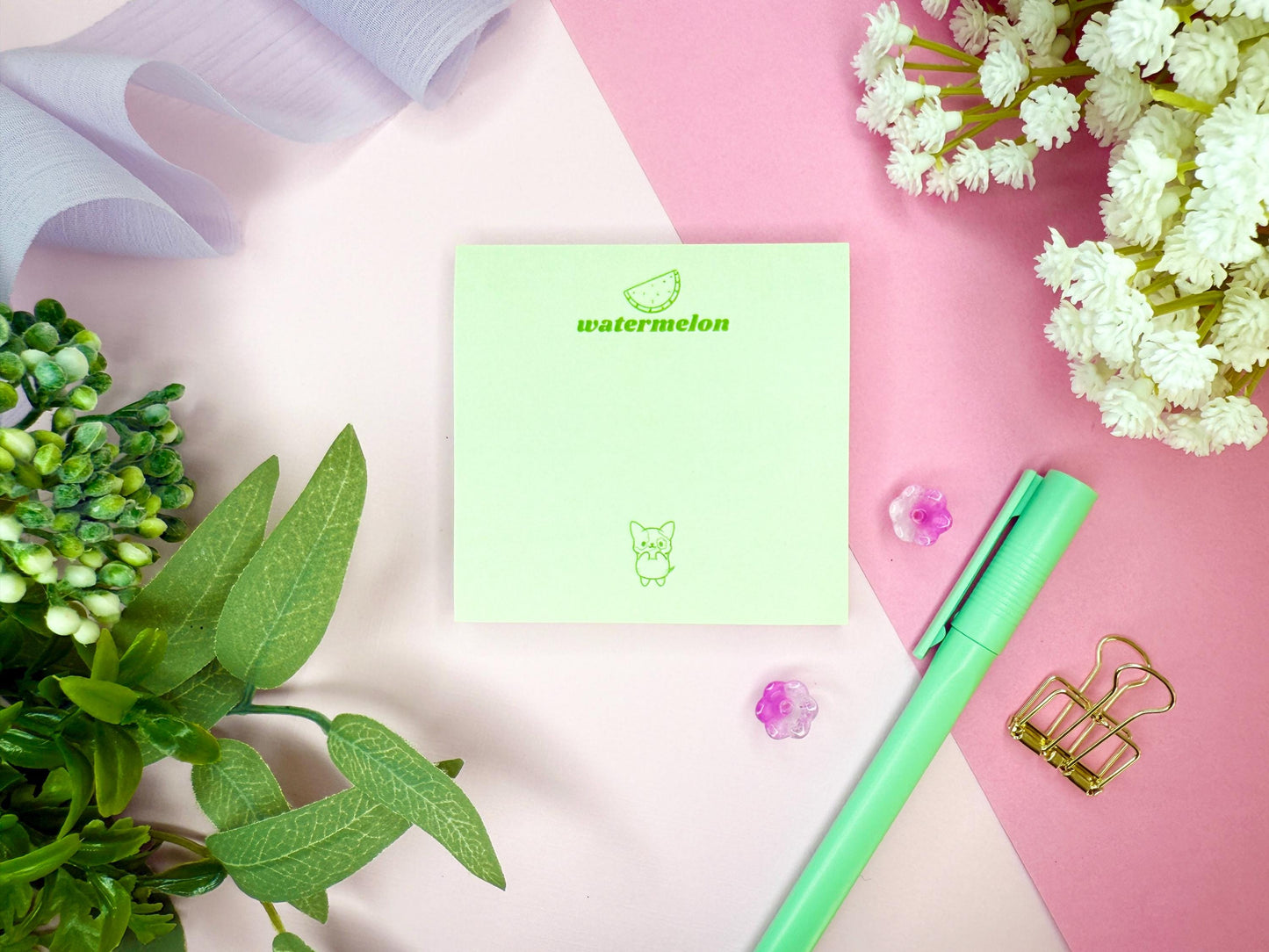 Light green notepad with "watermelon" branding and illustration as well as a kawaii dog with a green pen, and flowers on a pink and white background