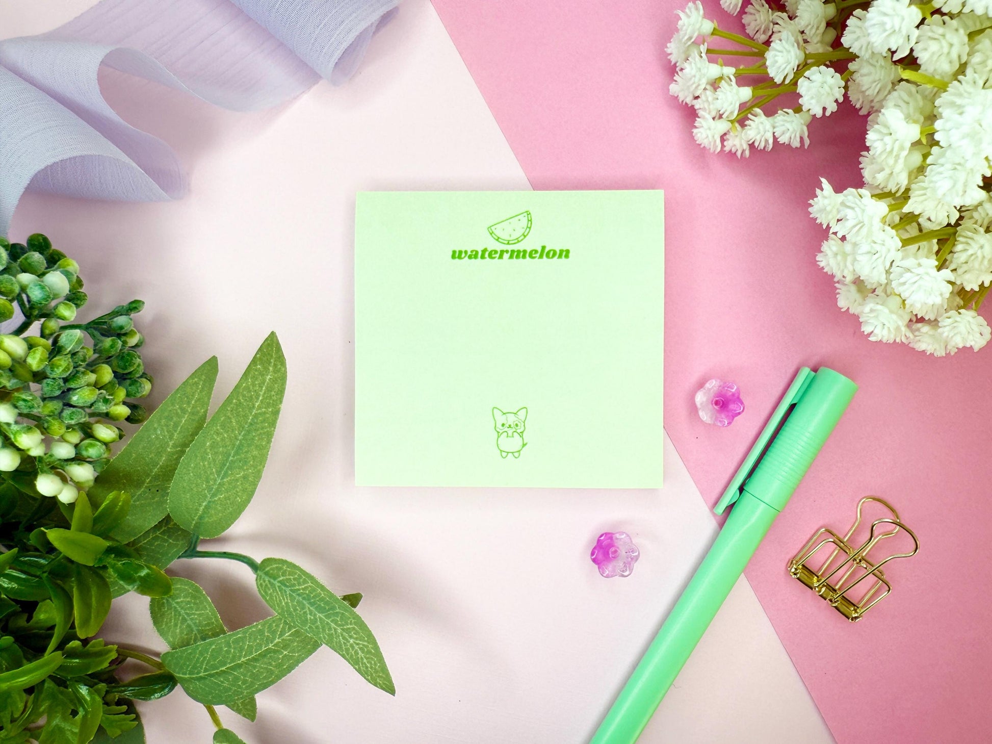 Light green notepad with "watermelon" branding and illustration as well as a kawaii dog with a green pen, and flowers on a pink and white background