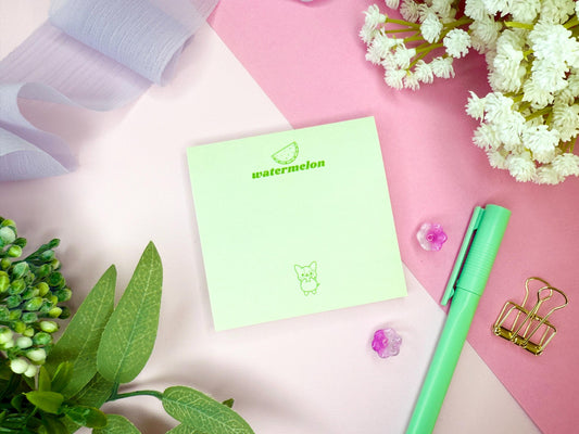 Light green notepad with "watermelon" branding and illustration as well as a kawaii dog with a green pen, and flowers on a pink and white background