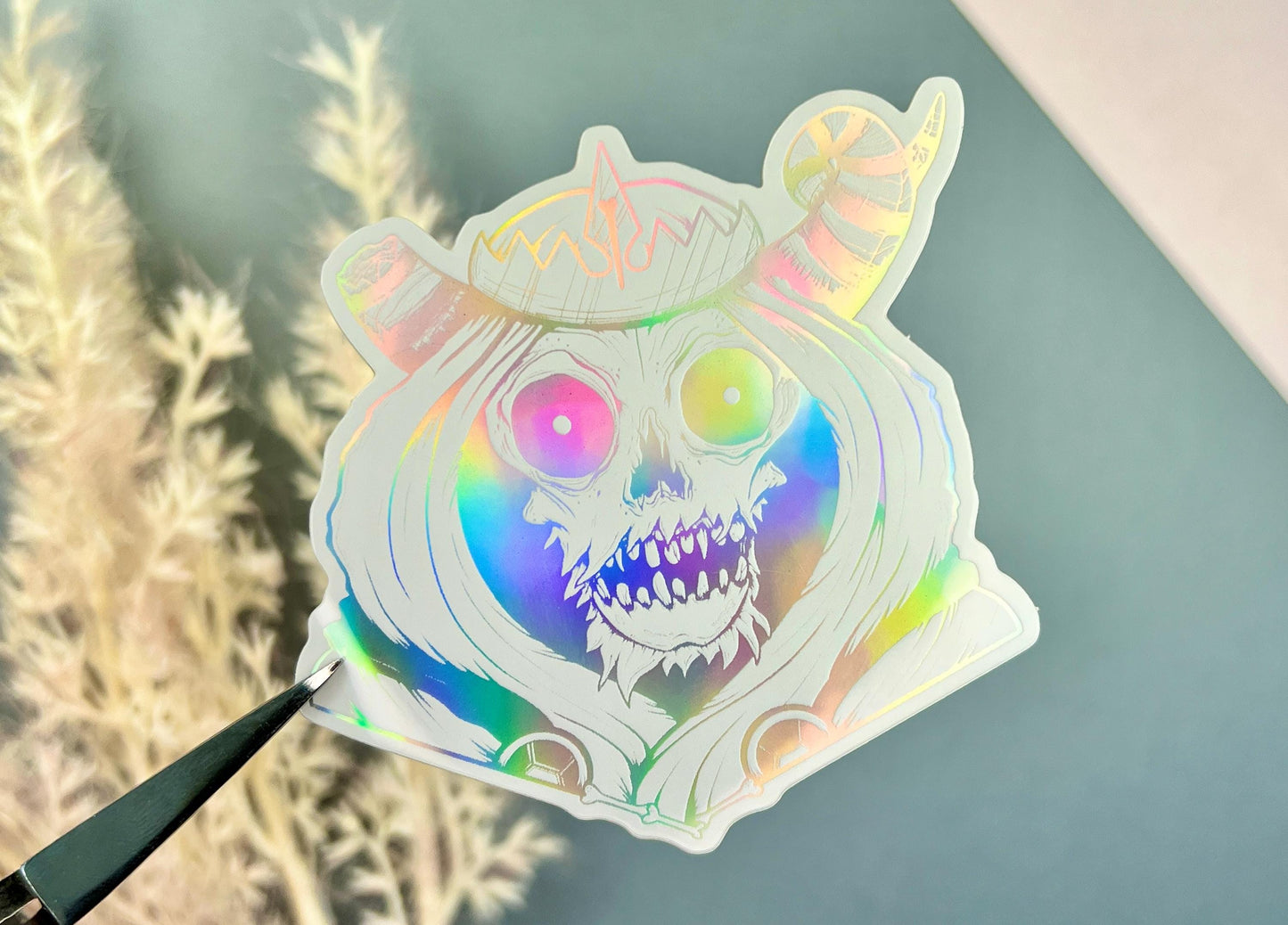 A white and silver holographic sticker of a skull with horns, a crown, a hood, and cape.