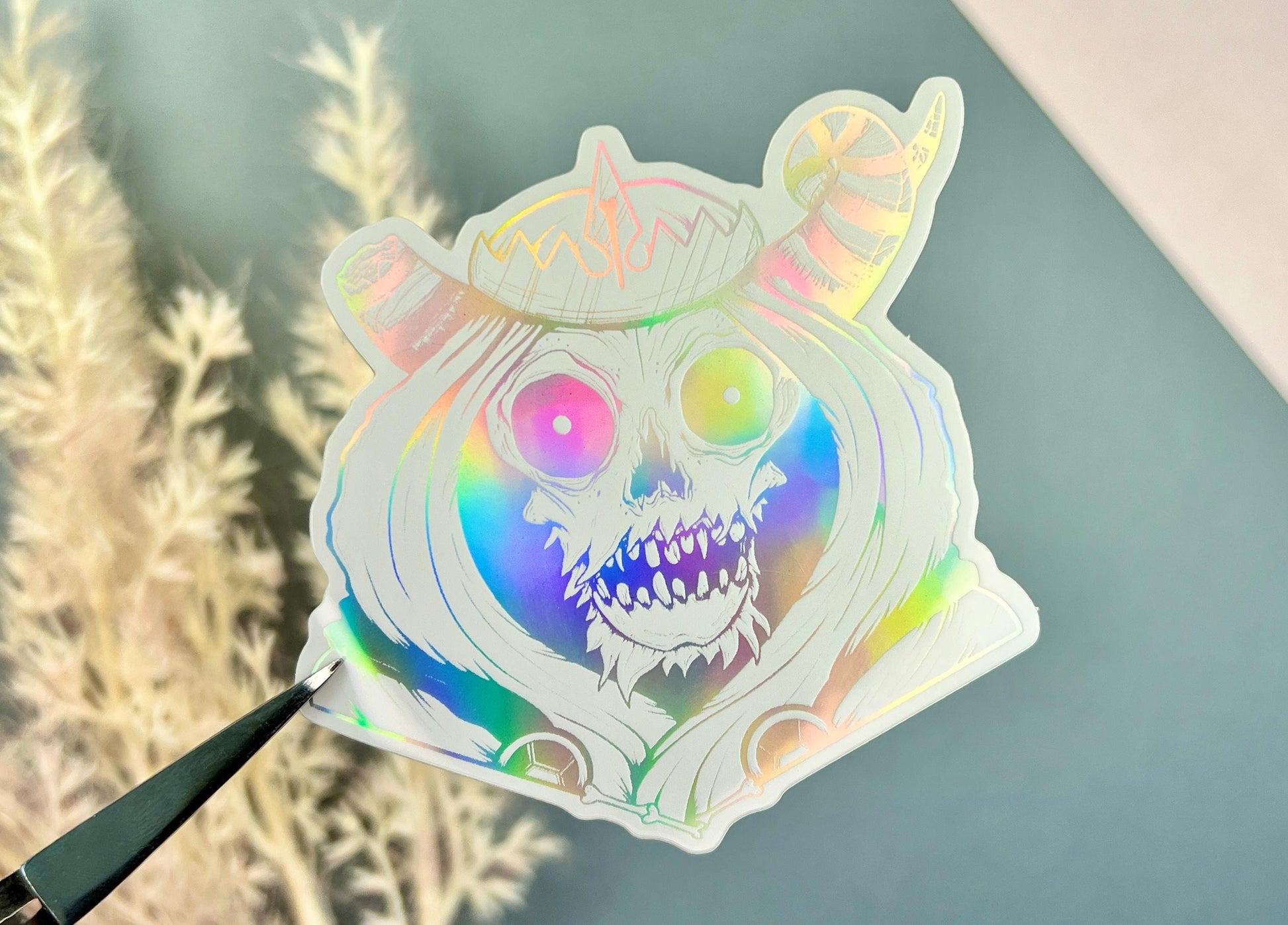 A white and silver holographic sticker of a skull with horns, a crown, a hood, and cape.