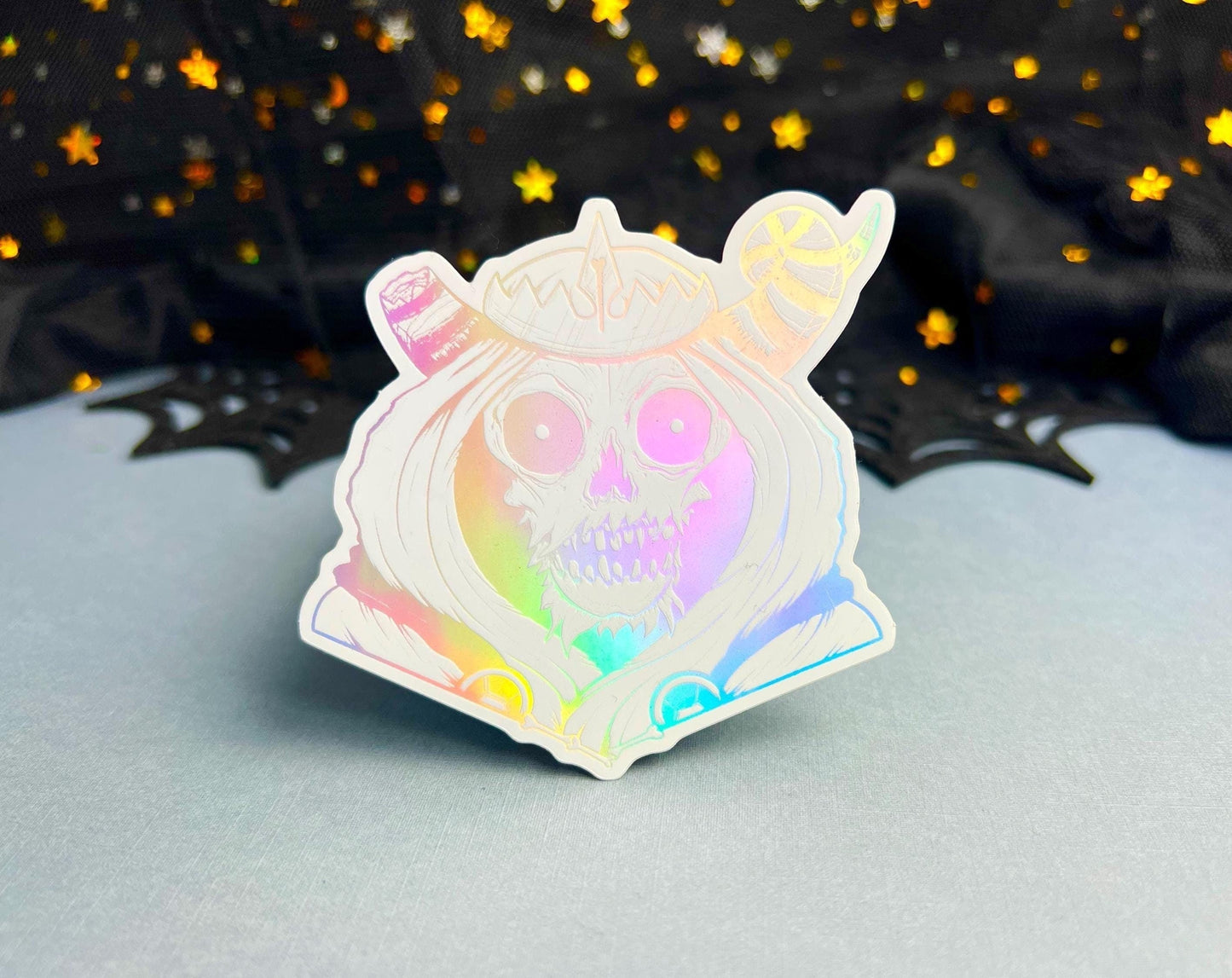 A white and silver holographic sticker of a skull with horns, a crown, a hood, and cape.