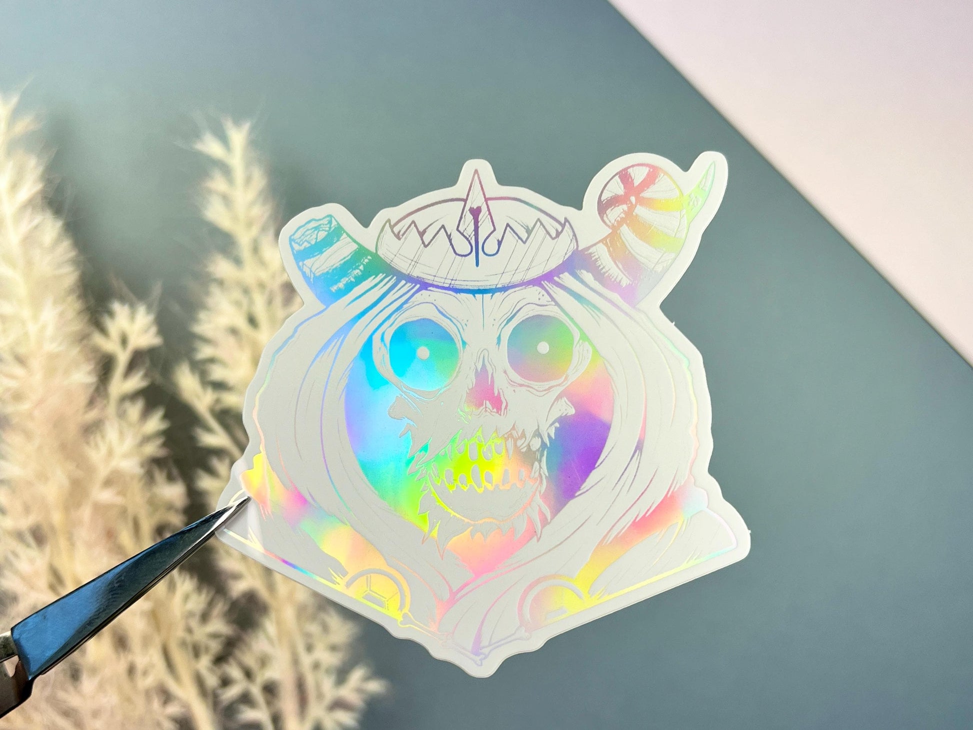 A white and silver holographic sticker of a skull with horns, a crown, a hood, and cape.