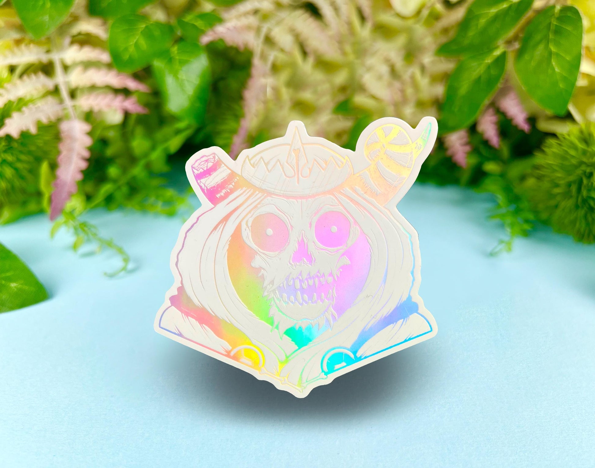 A white and silver holographic sticker of a skull with horns, a crown, a hood, and cape.