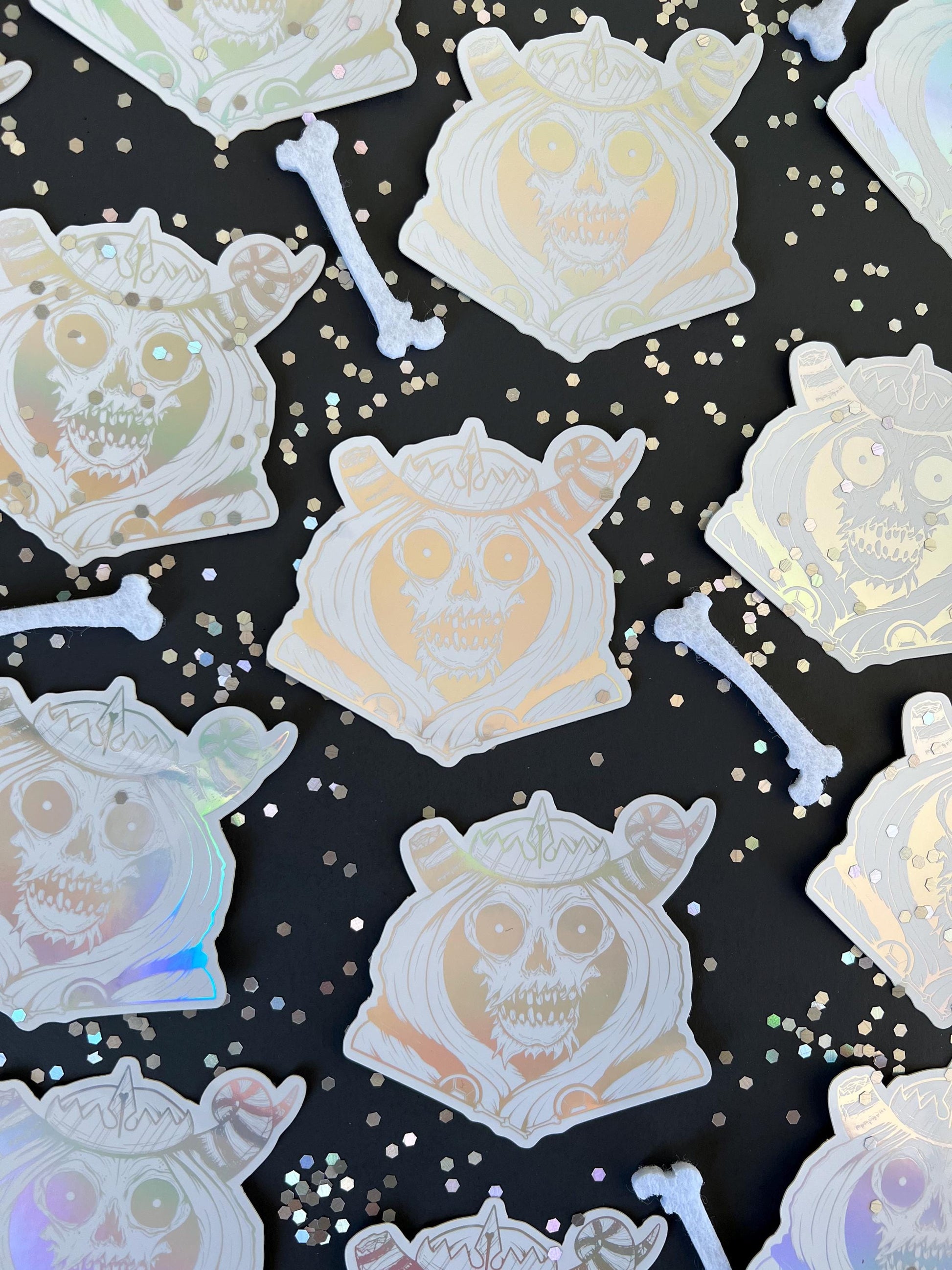 A white and silver holographic sticker of a skull with horns, a crown, a hood, and cape.