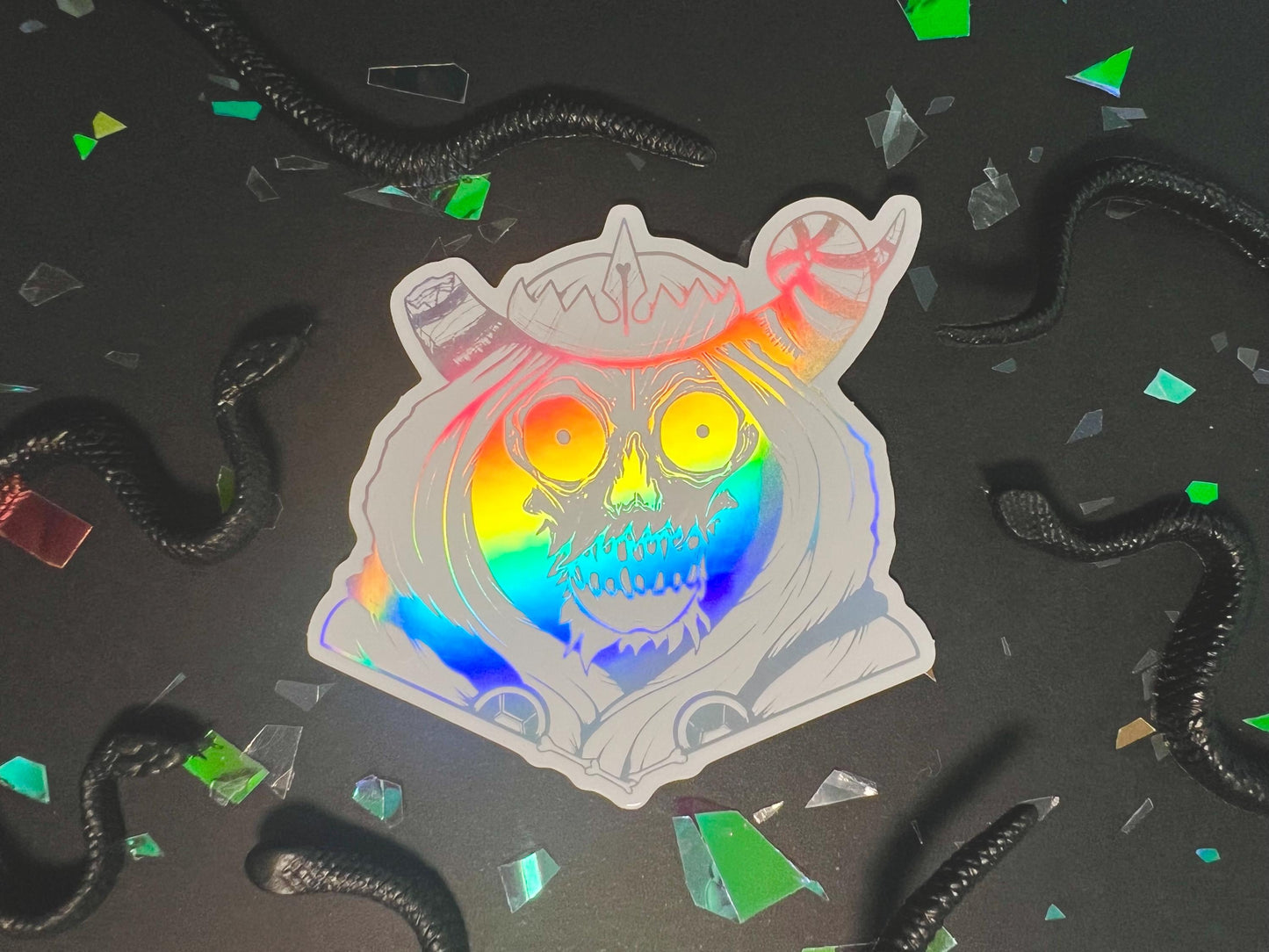 A white and silver holographic sticker of a skull with horns, a crown, a hood, and cape.