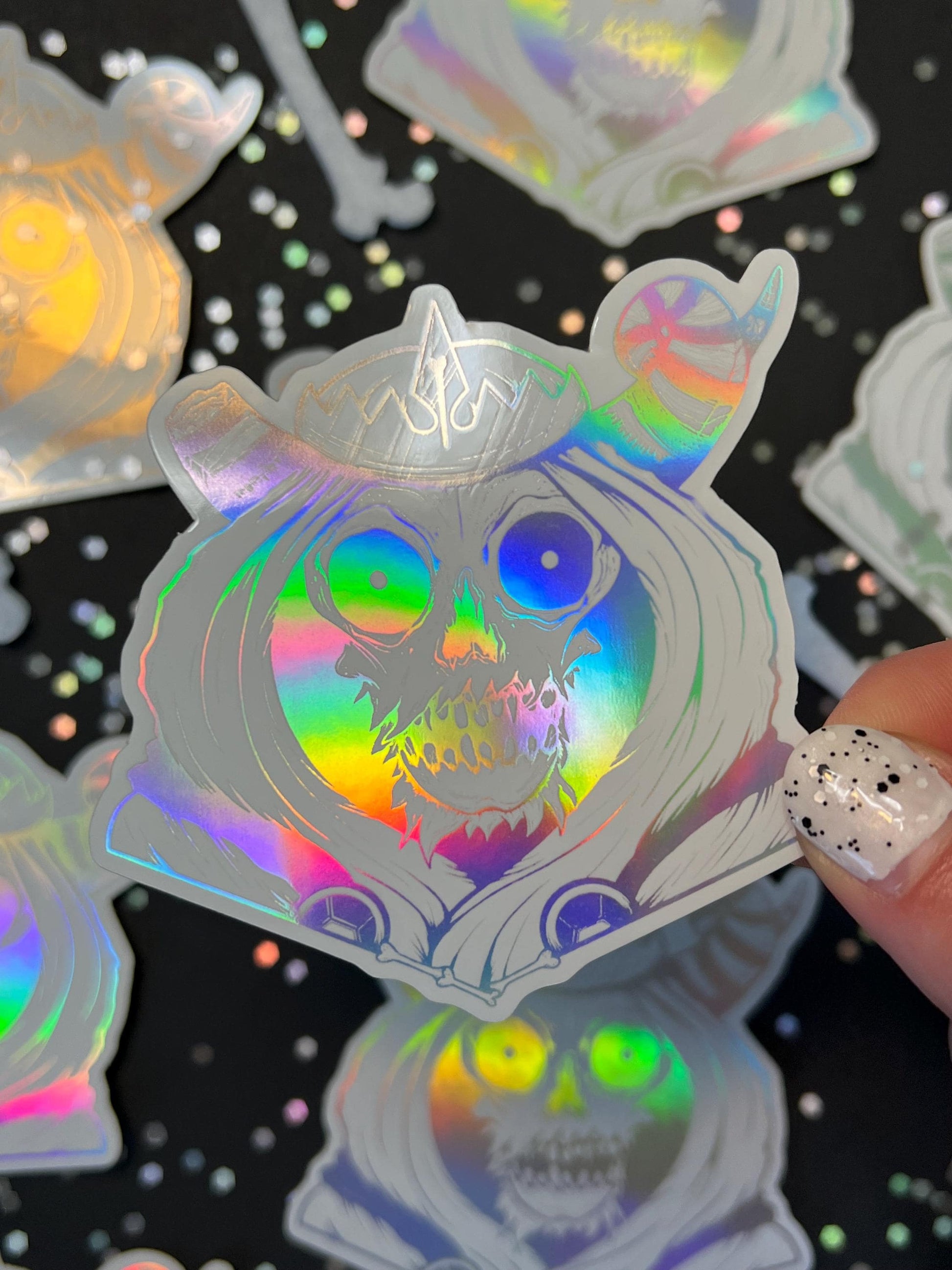 A white and silver holographic sticker of a skull with horns, a crown, a hood, and cape.